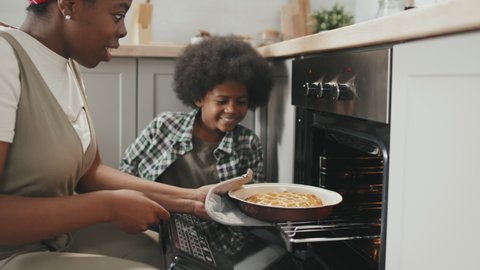 Slowmo shot of young African-American woman and her cute little son taking delicious homemade apple pie out of oven enjoying its taste
