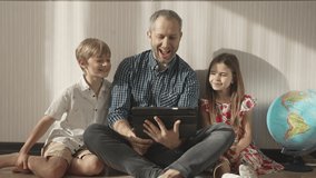 Family watching funny video by digital tablet and smiling. Father with children, boy and girl using portable device at home. Son, daughter and dad laughing to camera, enjoying time together at home.