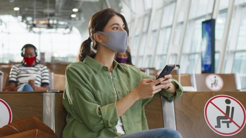 Charming young caucasian brown-haired girl wearing protective mask tapping and texting a message on the smartphone and sitting in the airport hall