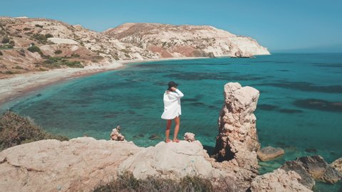 Hipster woman standing on Aphrodite Stone, back view. Female tourist on summer beach vacation. Beautiful waving white dress and hat. Cyprus sea coast. Adventure time. Nature landscape. Slow motion.