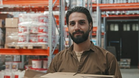 Slowmo tracking portrait shot of young man with beard holding boxes and smiling for camera at hardware store warehouse