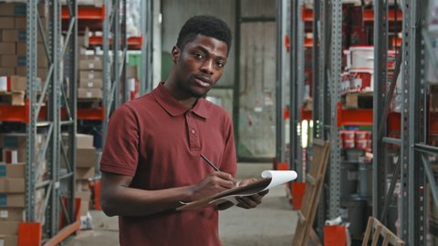 Slowmo tracking portrait of African-American male supervisor checking inventory and making notes on clipboard in warehouse, then turning towards camera and posing