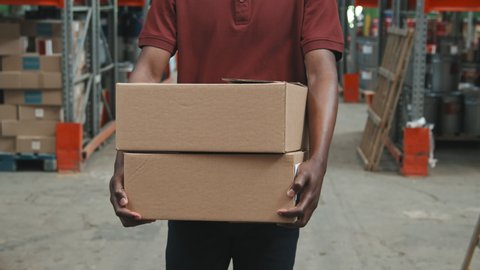 Slowmo tracking with mid-section of unrecognizable African-American worker carrying cardboard boxes and walking through warehouse