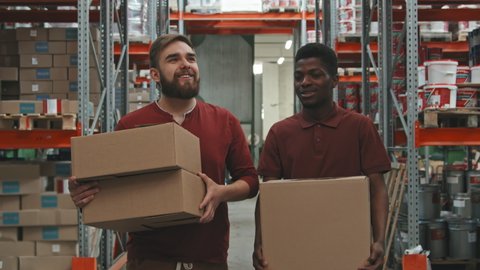 Slowmo tracking shot of cheerful male workers carrying cardboard boxes and chatting while walking through warehouse of hardware store