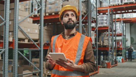 Handheld tracking with slowmo of bearded young male worker in reflective orange vest and hard hat typing on tablet and looking at items stacked on shelves in warehouse while stocktaking