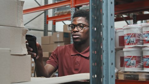 Handheld tracking with slowmo of African-American man in glasses using barcode reader and checking cardboard boxes in warehouse of hardware store