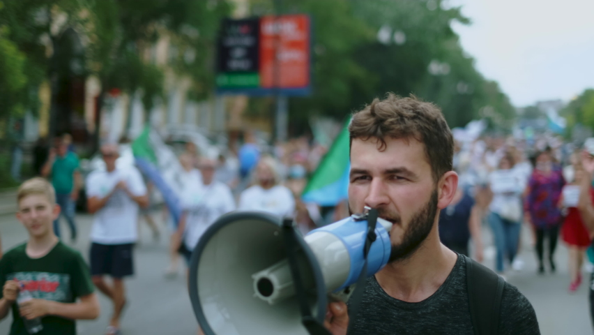 Angry political protester guy with bullhorn marches in protest crowd. Male rebel speaking on demonstration revolt resistance. Strike activist demonstrator man on opposition rally riot with megaphone. Royalty-Free Stock Footage #1077782291