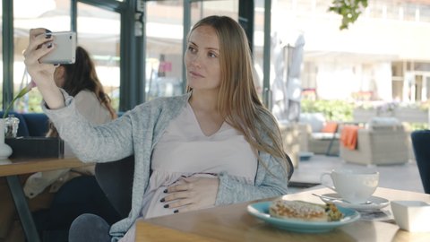 Joyful pregnant woman is taking selfie having fun in modern summer cafe sitting at table alone posing with sonogram image. Pregnancy and photos concept.