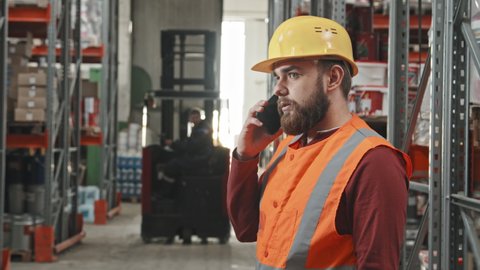 Slowmo tracking shot of bearded male supervisor in safety vest and hard hat talking on mobile phone in warehouse