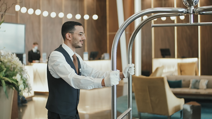 Medium shot of modern couple arrived in luxury hotel giving their luggage to young bellboy in uniform placing it on cart with other suitcases | Shutterstock HD Video #1077785747