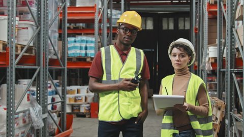 Handheld slowmo tracking of African-American man and Caucasian woman wearing hard hats and vests and holding clipboard and walkie-talkie while discussing work in warehouse, then posing