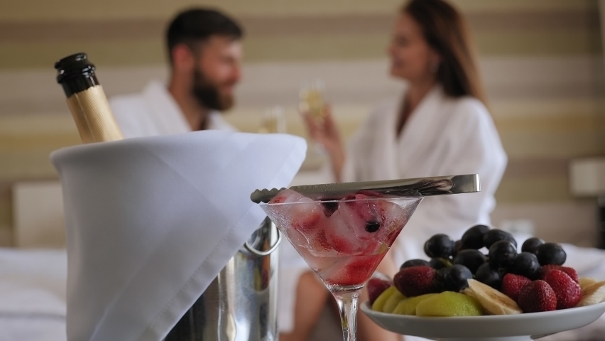 Close-up of a bucket for champagne, ice and fruits on a table in a hotel, in the background a loving couple is sitting on the bed. Blurred background. Royalty-Free Stock Footage #1077786983