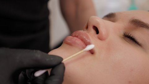 Close-up cosmetologist moistens the lips of a young woman with cream. Permanent makeup procedure in a modern beauty salon.