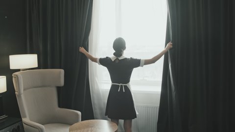 Zoom-out rear-view slowmo shot of female housekeeper opening black curtains in luxury hotel room