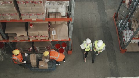 Top view shot of workers in safety vests and hard hats working in warehouse with cardboard boxes stacked on shelves