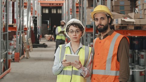 Slowmo tracking portrait shot of female manager with tablet and her male colleague wearing safety vests and hard hats and posing for camera in busy warehouse