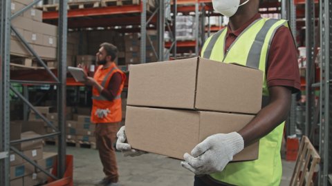 Slowmo mid-section tracking of unrecognizable African-American male worker in face mask, safety vest and gloves holding cardboard boxes in warehouse. Male supervisor stocktaking in background