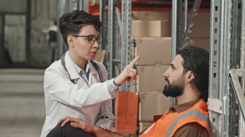 Handheld slowmo tilt up of female doctor in white coat examining male warehouse worker resting by shelves in workplace