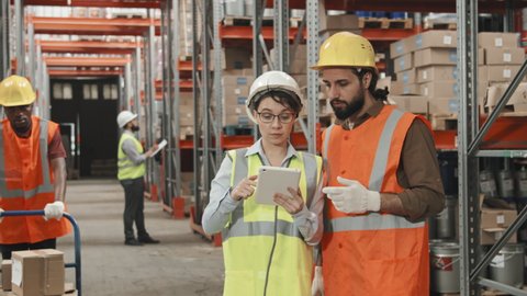 Slowmo tracking shot of female manager in hard hat and vest showing something on tablet and talking to male worker in warehouse while African-American male worker transporting products on cart
