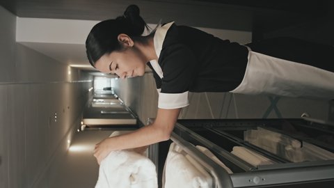 Vertical shot of young female housekeeper in uniform taking fresh white towels from hand cart, opening hotel room with card key and walking inside for cleaning