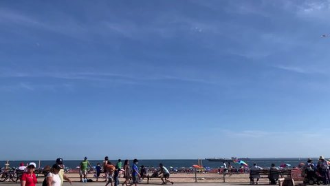 Brooklyn, NY, USA, 8.15.21 - The view of Coney Island from the boardwalk. There are crowds of people walking along the boardwalk and enjoying the sand beaches. A large cargo boat is floating along too