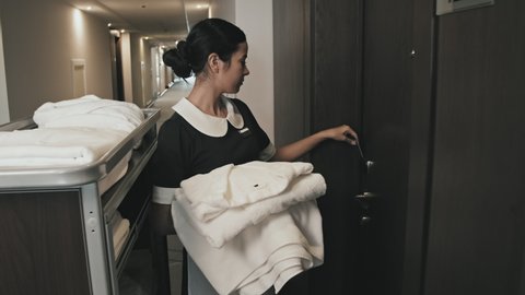 Medium shot of female housekeeper with towels, linens and cleaning supplies on hand cart walking along long corridor, entering hotel room for cleaning opening door with card key