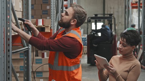 Slowmo handheld shot of female manager with tablet talking to bearded male worker scanning barcodes of boxes stacked on shelves in warehouse
