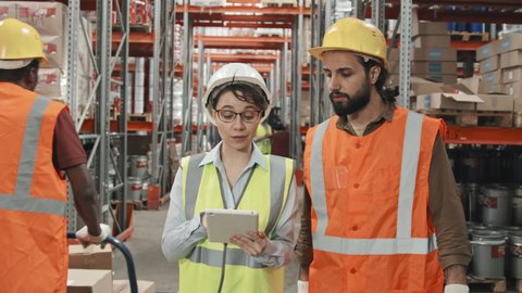 Slowmo tracking of female supervisor in safety vest and hard hat looking at tablet and talking to male colleague while walking through busy warehouse