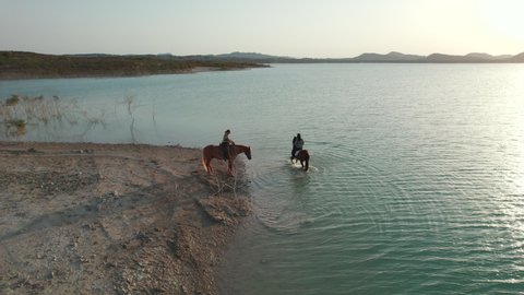 Aerial drone point of view two young adult caucasian women rides on horses outdoor in the Embalse de La Pedrera lake view, Orihuela. Spain. Concept of hobby, equestrian sport and active lifestyle