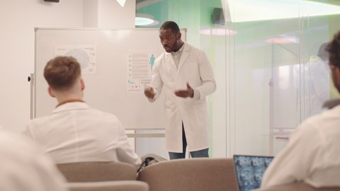 Medium shot of young African-American male university professor teaching Anatomy to group of medical students in modern classroom with posters of human brain on whiteboard