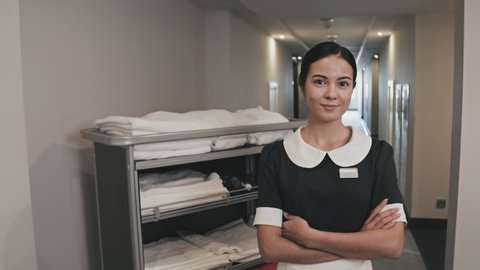 Medium slowmo portrait of young pretty female housekeeper standing with hands folded in hotel corridor smiling to camera