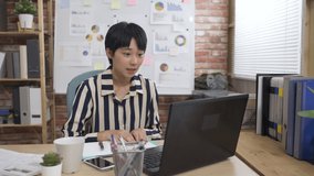 sincere asian female is using hand gestures and writing notes while communicating with her client through video conference call on laptop in the office.