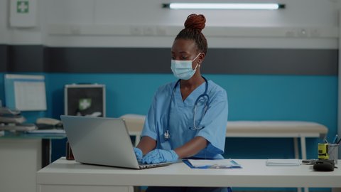 Portrait of medical assistant with uniform and face mask in cabinet at healthcare facility. Young nurse sitting at desk working on laptop in office during coronavirus pandemic