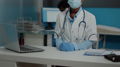Doctor of african american ethnicity sitting at desk with laptop and documents while wearing face mask and white coat looking at camera. Black woman working as medic in healthcare cabinet