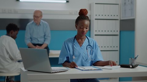 Young adult nurse sitting at desk with laptop and documents in examination hospital room. Medical assistant with stethoscope looking at camera while working at healthcare clinic