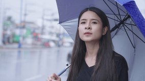 A beautiful east asian woman stands alone on roadside waiting for taxi or bus in hurry raining time, holding umbrella in rain, hurry rush hour, tropical climate change, urban style in rainy season