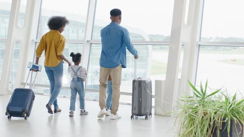 Back view on the multiracial family - mother, father, daughter and son - walking through the airport hall and carrying their suitcases on the wheels before the departure to the vacation