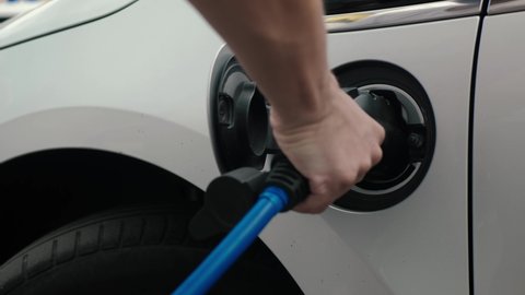 Male hand inserts power connector into EV car. Unrecognizable man plugging in charging cable to to electric vehicle and charges batteries