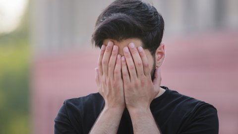 Scared upset middle eastern bearded young man wearing black t-shirt cover face feel frightened, fearful Indian male student close eyes afraid or terrified avoid seeing bad, peeping through fingers