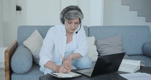 Elderly mature old focused woman wearing headset, studying with teacher online, learning foreign language, making notes in notebook. Internet distant education from home is available at older age