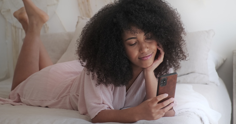 A peaceful black woman holding a smartphone and texting while lying in a cozy bed, a smiling woman using smartphone online services, browsing social media, shopping, purchasing online in a mobile app Royalty-Free Stock Footage #1077821186
