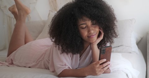 A peaceful black woman holding a smartphone and texting while lying in a cozy bed, a smiling woman using smartphone online services, browsing social media, shopping, purchasing online in a mobile app