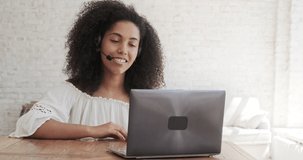 Black lady wearing a headset begins a lesson online, looks at a laptop screen, explains to a student, learning with an educator, business work from home employing a teleconference software approach.