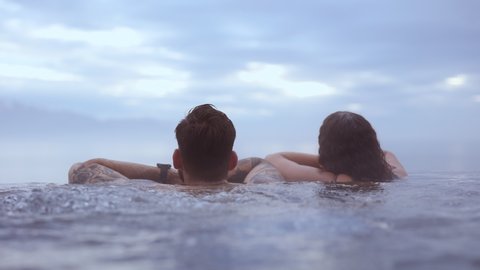 REYKJAVIK, ICELAND - JUNE 2021: Beautiful steady shot of a couple bathing in an infinity pool with the landscape of the majestic blue skies with fluffy clouds, with the camera placed at an angle