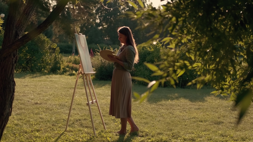 Creative woman artist paints a picture with a brush while standing near the easel in nature. Woman Artist, Art for Sales, Inspiration in Nature. Royalty-Free Stock Footage #1077836834