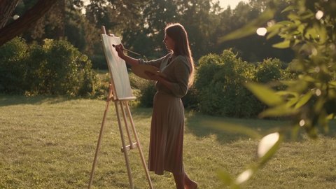 Creative woman artist paints a picture with a brush while standing near the easel in nature. Woman Artist, Art for Sales, Inspiration in Nature. วิดีโอสต็อก