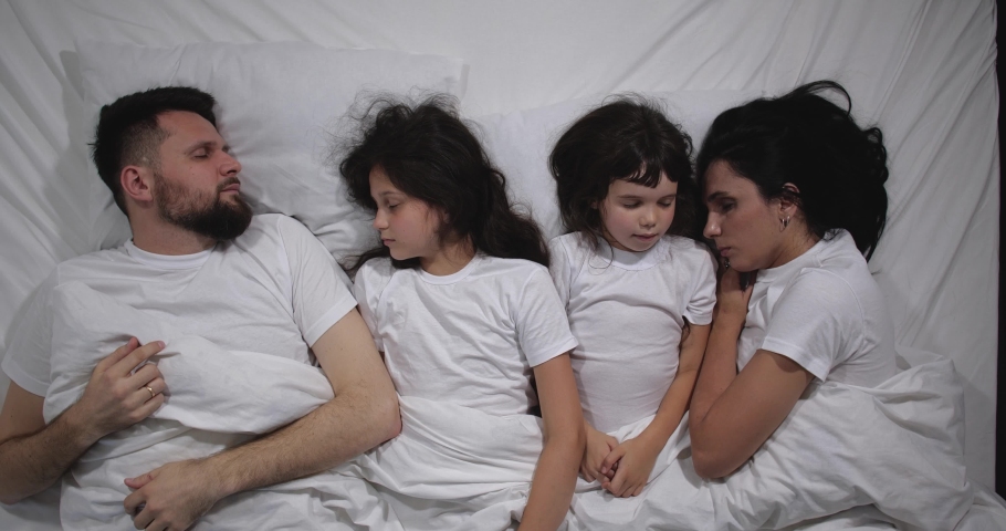 Top view of caucasian family with two children waking up in bed with white linens in light room. High angle shooting. A family of four gets out of bed. Children wake up their parents