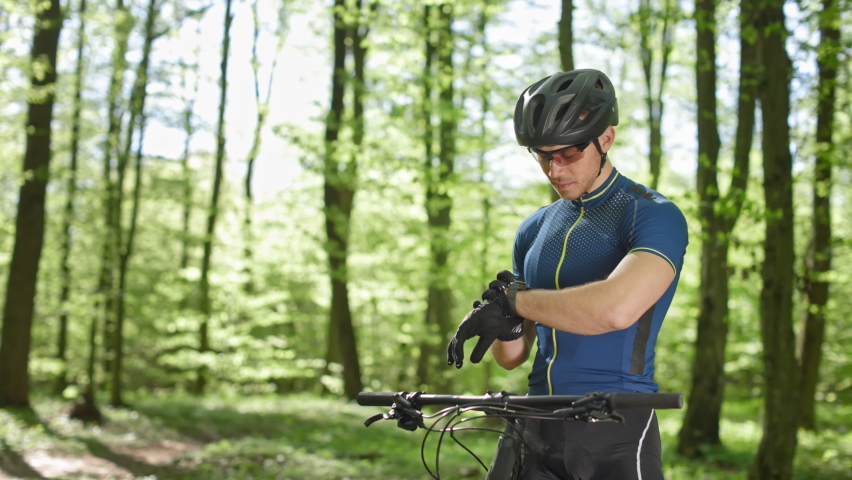 The cyclist is viewing the training data on the watch. He is standing next to an MTB bike in the woods. He's wearing cycling clothes. The camera is moving from left to right. 4K Royalty-Free Stock Footage #1077839243
