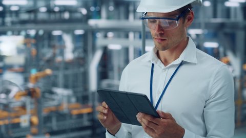 Male Engineer in Safety Goggles and Hard Hat Using Tablet Computer and Looking Out of the Office at a Car Assembly Plant. Industrial Specialist Working on Vehicle Production in Technological Factory.