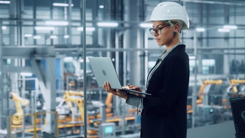 Female Engineer Wearing White Hard Hat, Using Laptop Computer and Looking Out of the Office at a Car Assembly Plant. Industrial Specialist Working on Vehicle Design in Technological Facility.